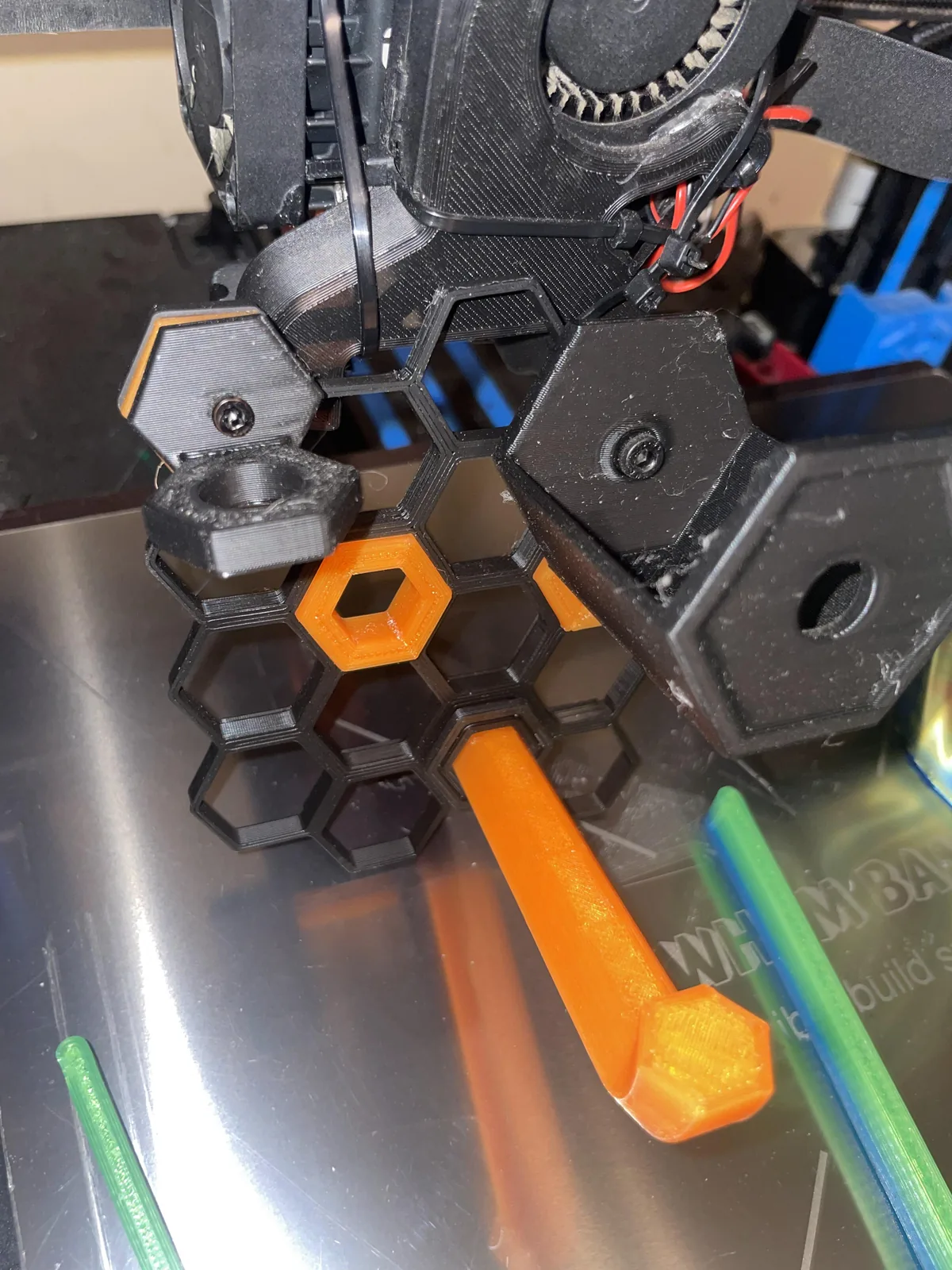 A close view of a printer toolhead with black printed hexagonal parts and an orange handle on a steel build plate