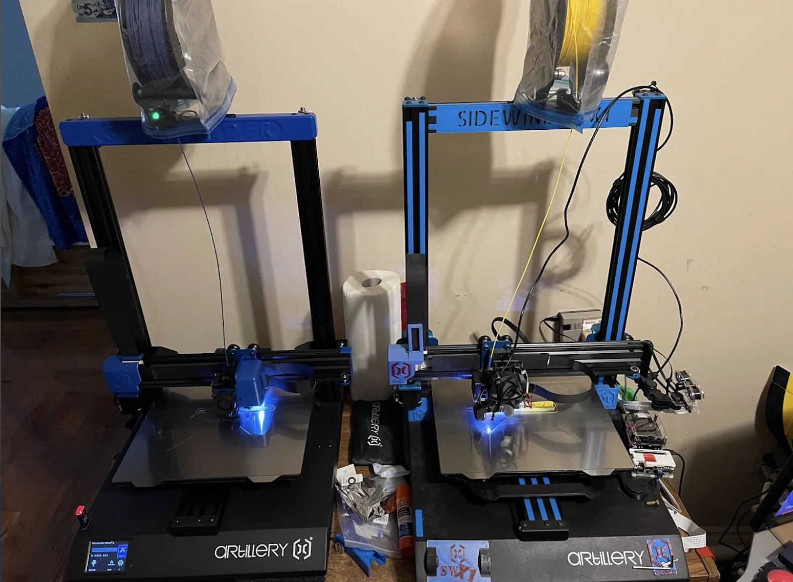 Two blue and black FDM 3D printers side by side in a home shop, both mid-print with blue glowing hotends