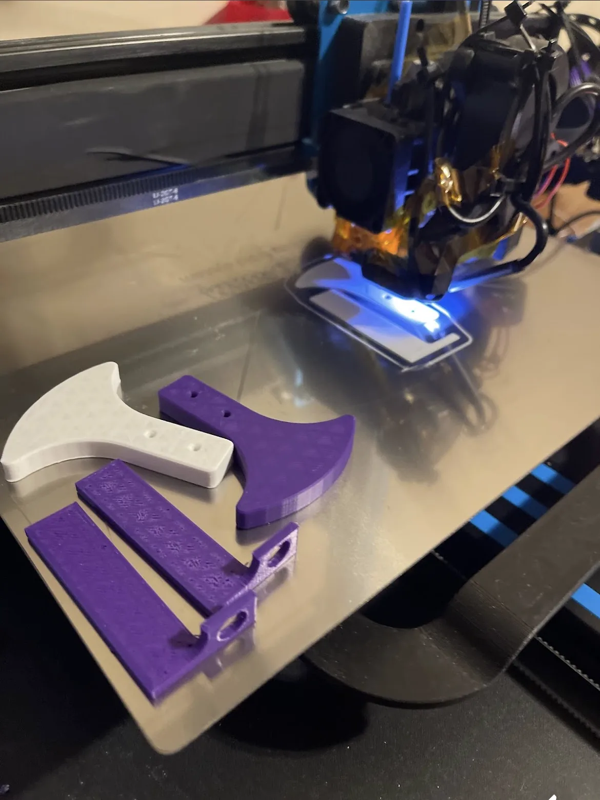 Purple and white printed brackets and flat parts on a print plate beneath a printer toolhead