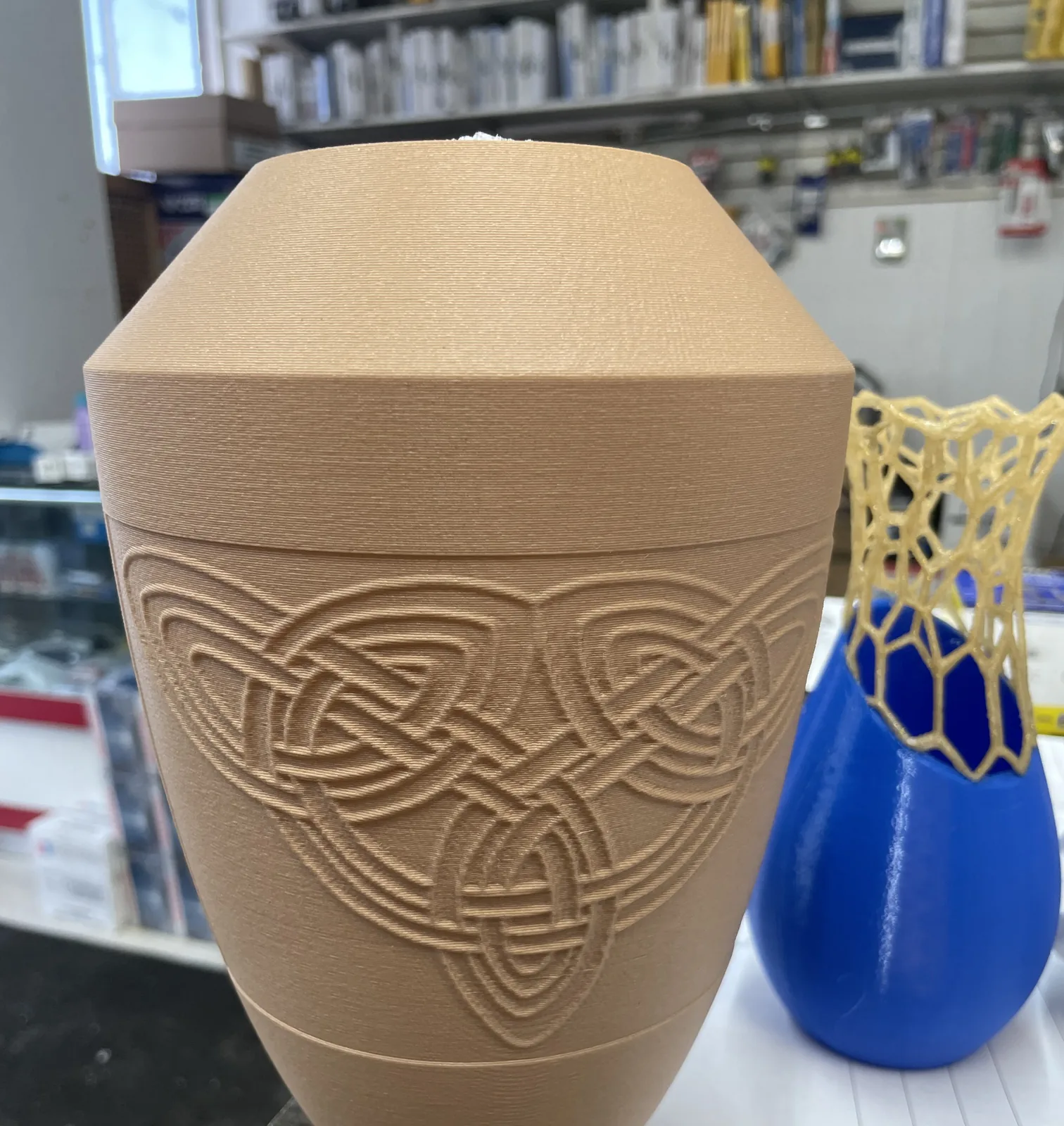 A tan printed lidded vessel with an interlaced knot relief pattern beside a blue vase with a yellow lattice neck