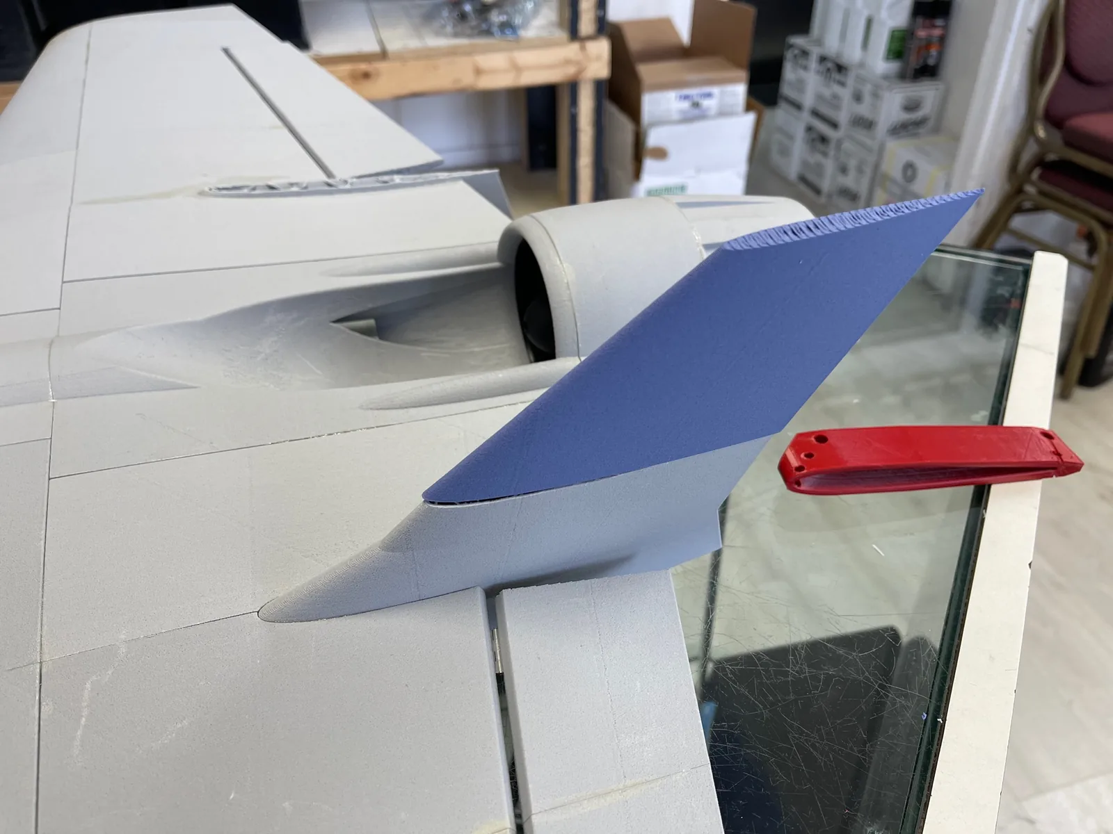 A grey printed jet wing and ducted-fan housing with a blue printed vertical fin and a red printed fitting resting on a glass table edge