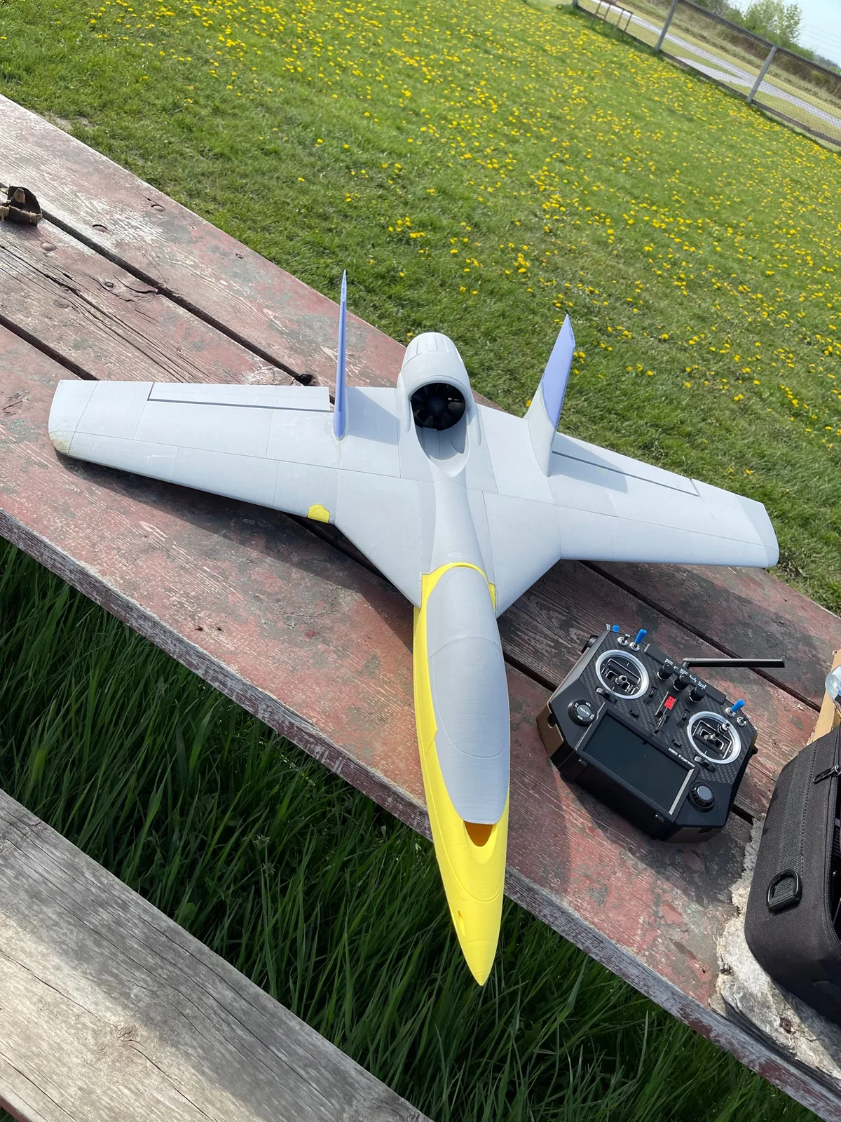 A grey R/C jet with a yellow nose and blue fins on a wooden picnic table beside a black radio transmitter
