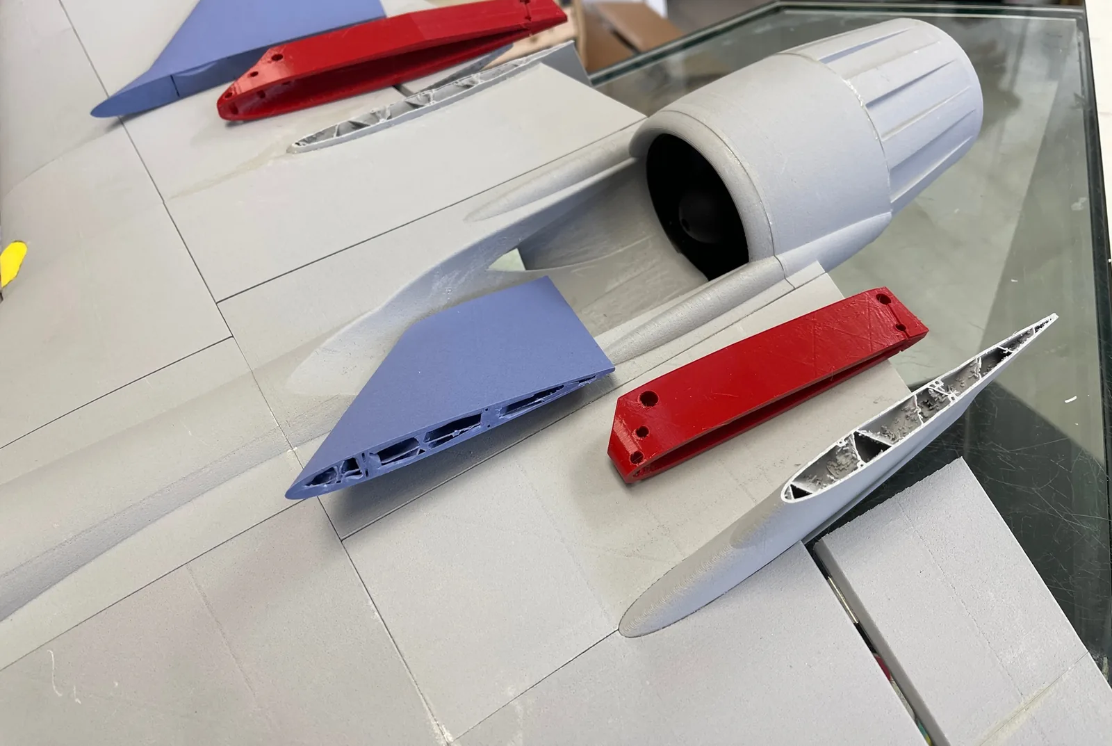 Printed jet parts laid on glass: blue fin, red brackets, grey ducted-fan housing and a cut-away fin