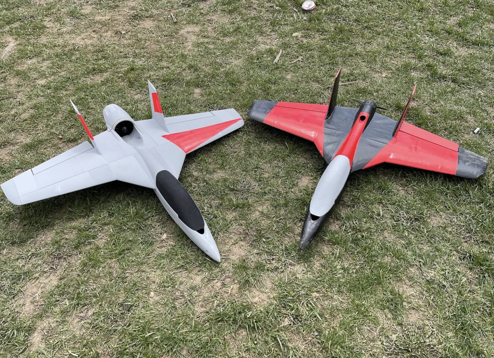 Two 3D-printed R/C jets lying on grass, one grey with a black canopy and red tips, one grey and red with a dark nose